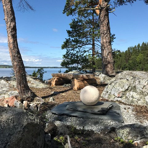 En bänk, konstverk (Tystnadspärla i granit) och träd, i bakgrunden vatten. Foto.