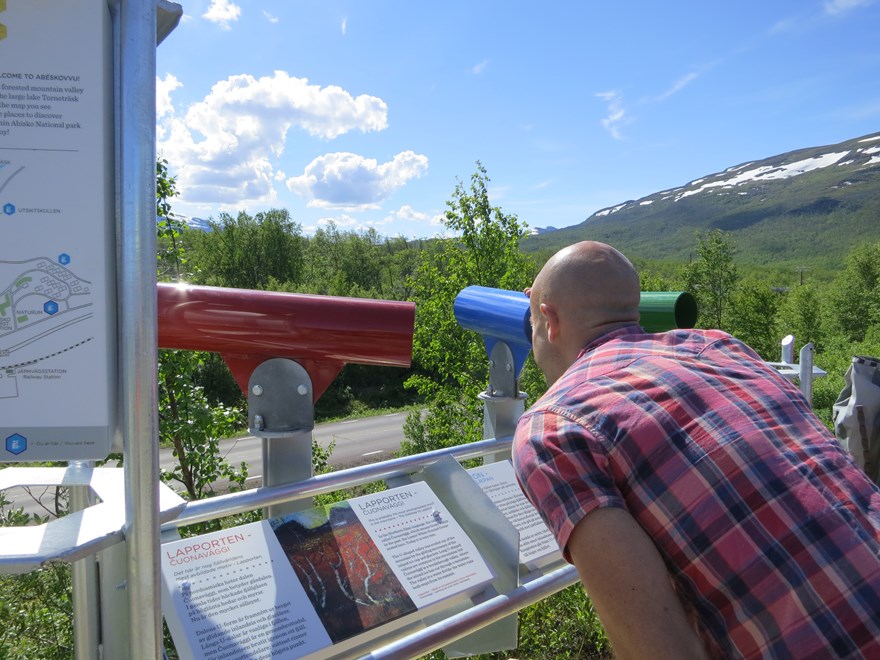 A person looks through binoculars at an information sign. In the background mountains with snow. Photo.