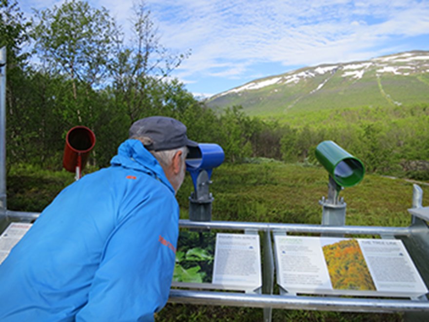 En person kollar i en kikare vid en informationsskylt. I bakgrunden berg med snö. Foto.