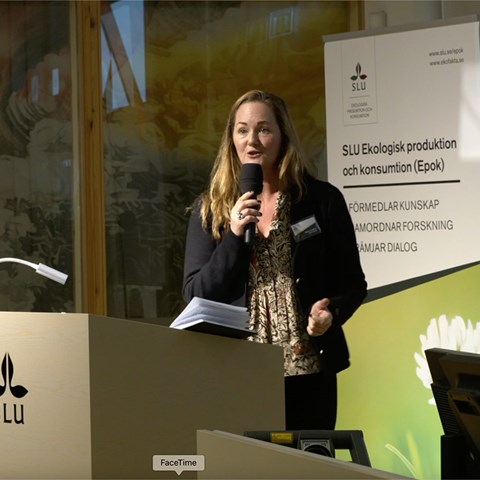 Maria Tunberg står vid en talarstol och pratar i en mikrofon. Bakom henne en grön rollup med texten Epok.