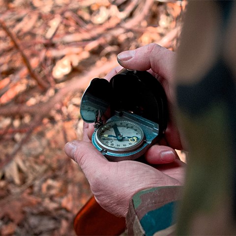 Två händer håller en kompass, i bakgrunden syns skogsmark. Foto.