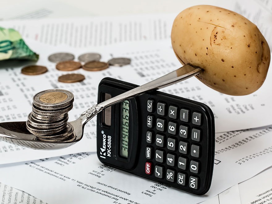 A spoon holding coins at one end and a potatoe at the other end balances on a calculator. Photo.