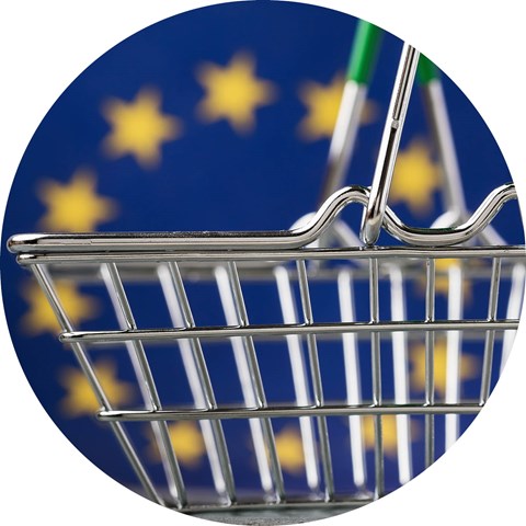 En kundkorg i miniatyr framför en EU-flagga. Foto.