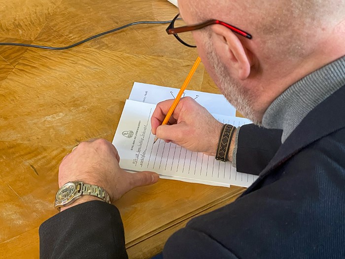 En man skriver på ett papper vid ett bord. Foto.