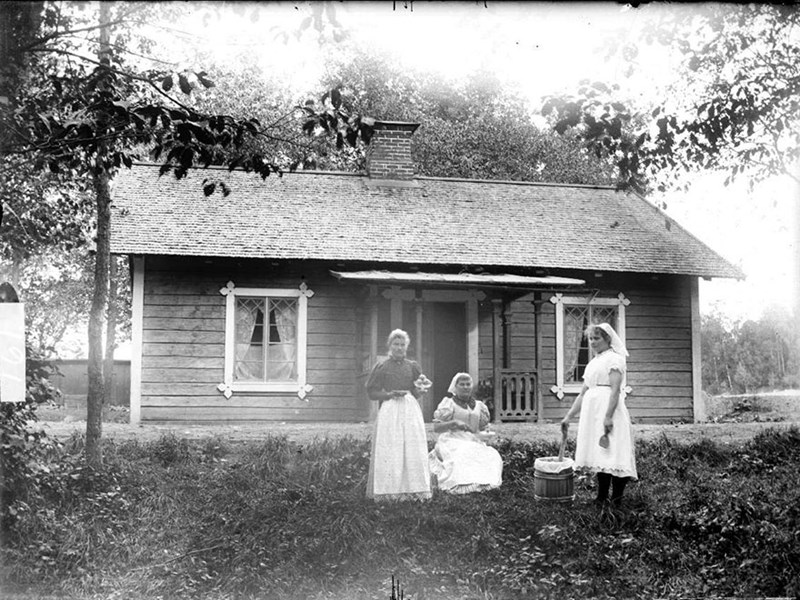 Gammalt svartvitt fotografi med tre kvinnor utanför en mindre träbyggnad.