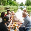 Ett sällskap med gamla och unga personer har picknick vid en å. Foto.