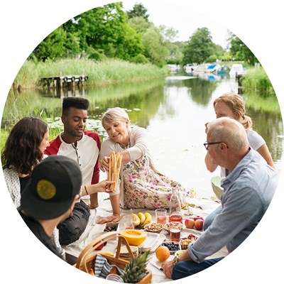 Ett sällskap med gamla och unga personer har picknick vid en å. Foto.