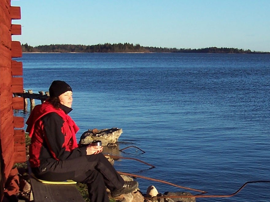 En kvinna sitter invid en röd husvägg med utsikt över havet. Foto.