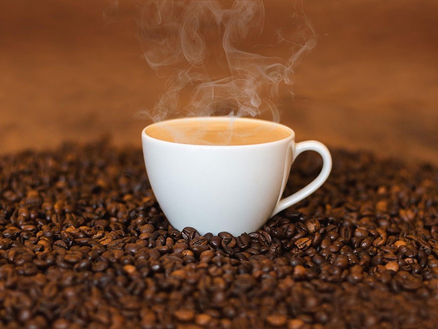 En rykande kopp kaffe står omgiven av kaffebönor. Foto.