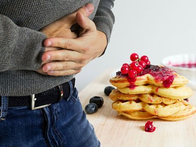En man håller sig för magen som om han har magknip. Bakom honom syns en hög med pannkakor med sylt. Foto.