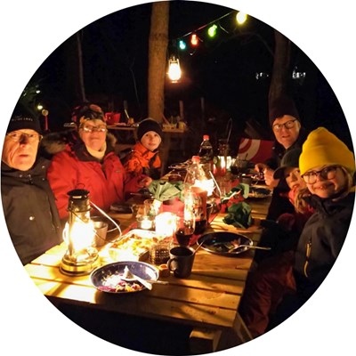 Sex personer sitter vinterklädda vid en bord med mat på. Det är mörkt ute och ljus hänger i träden. Foto.