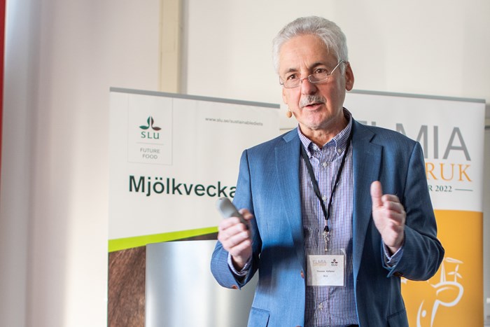 En man med grått hår står och pratar och gestikulerar med händerna. Foto.