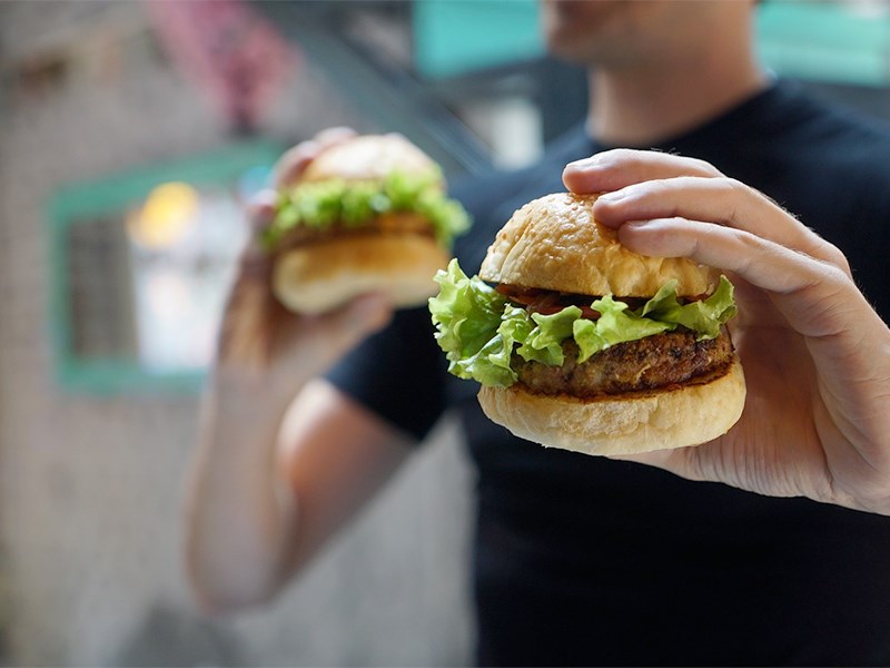 En person håller hamburgare i varje hand utomhus. Foto.