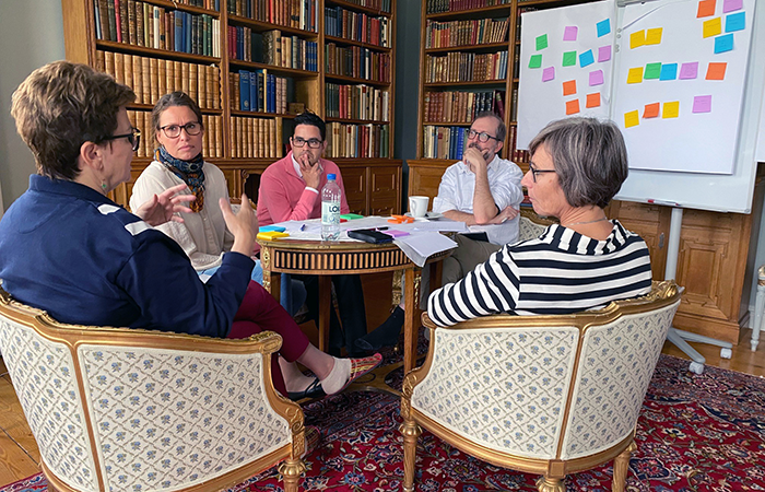 Three women and two men sit on a couch and two armchairs in a mansion. They discuss and use post-it notes and flipcharts. Photo.