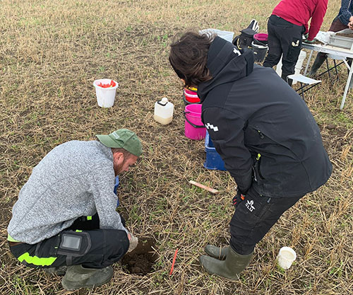 A man is squatting and digging in the ground, a woman stands next to him and watches. In the background, two people are seen next to a table sampling soil in a field. Photo.