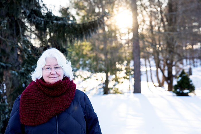 A woman with white hair and a big burgundy scarf in a forest covered in snow. Photo.