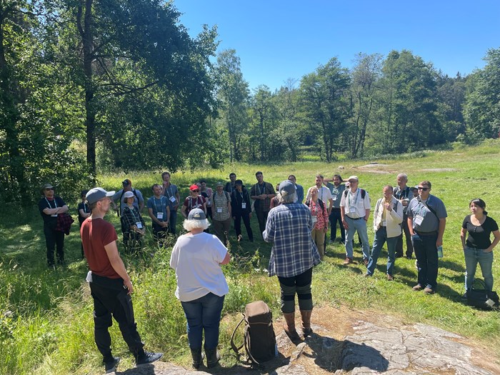 Three people stand in front of a large group of people next to a forest. Photo.