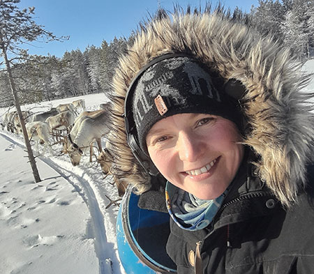 A young woman in close-up wearing warm clothes, behind her you can see a reindeer herd and there is lots of snow and a clear blue sky. Photo.