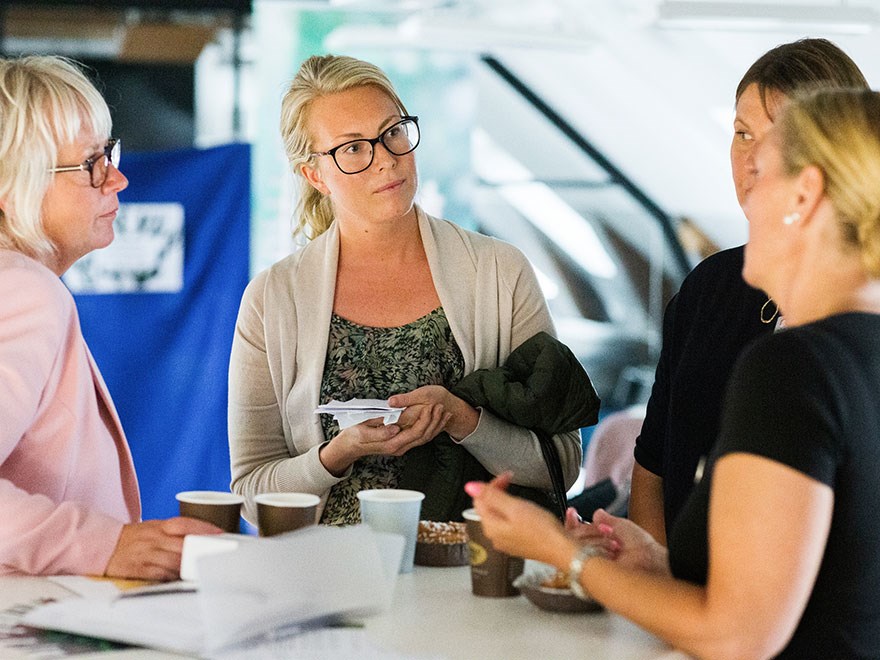 Four women are standing and talking around a table. Photo.