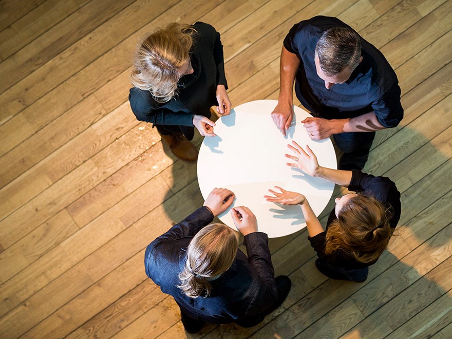 Three people are standing at a round table. Photo.