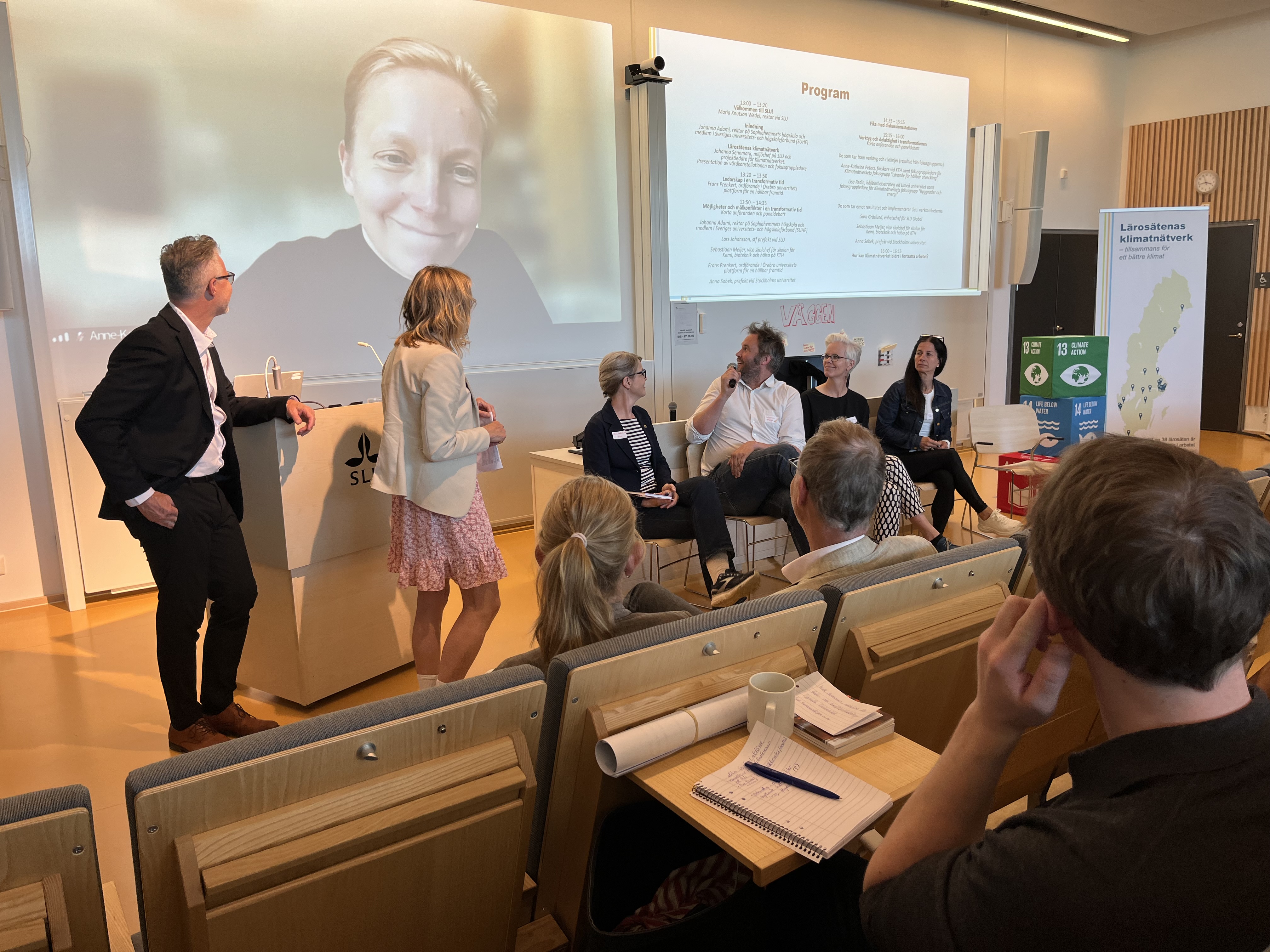 ett panelsamtal, två personer står och fyra sitter ned. de vänder sig mot en kvinna som är med via länk