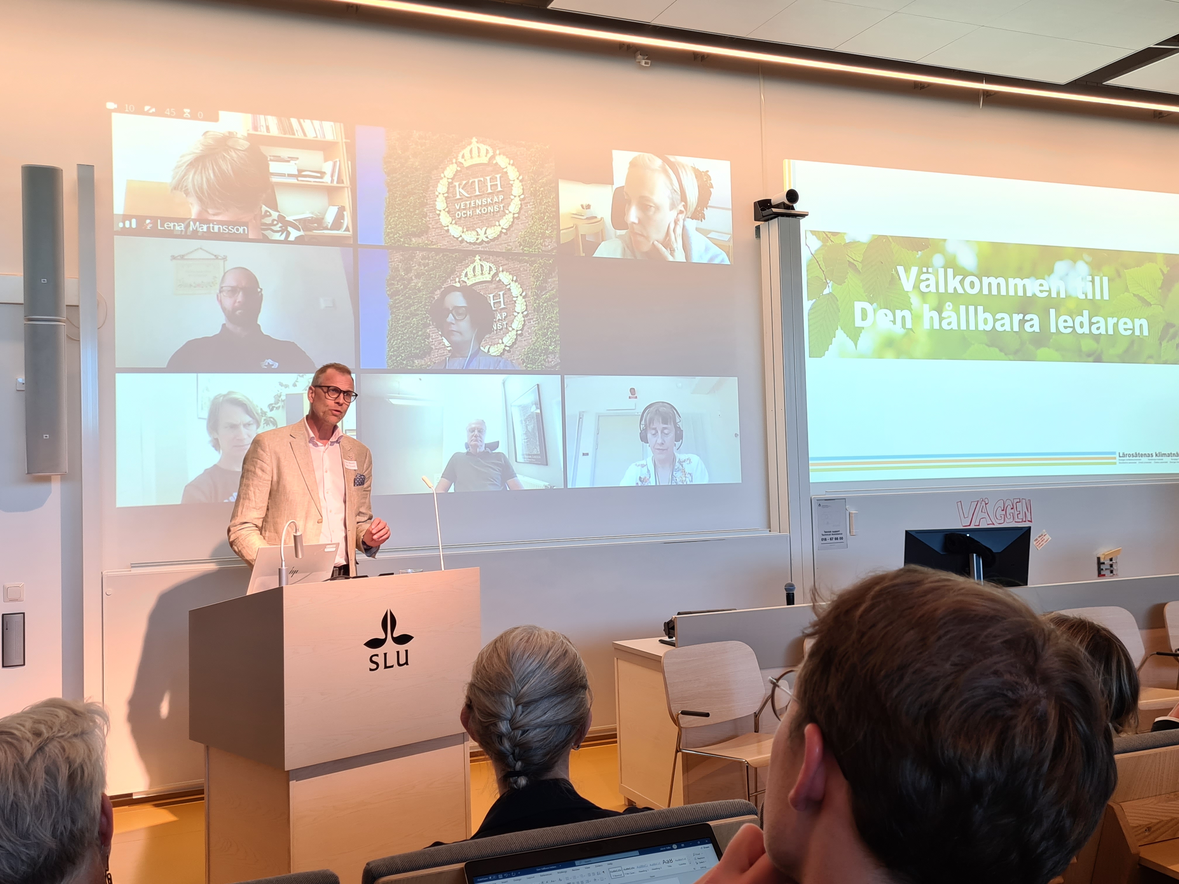 En man i ljus blazer står i talarstolen. Bakom honom bilder från deltagare i zoomlänk.