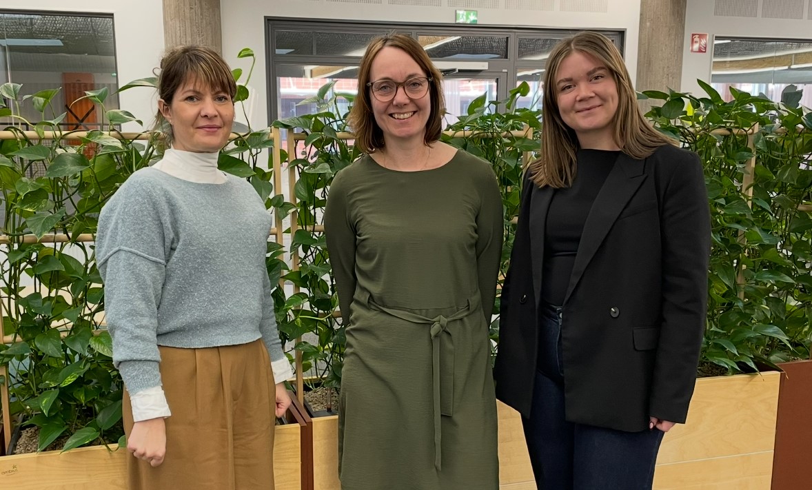 Tre kvinnor står tillsammans framför en rumsavskiljare med gröna växter