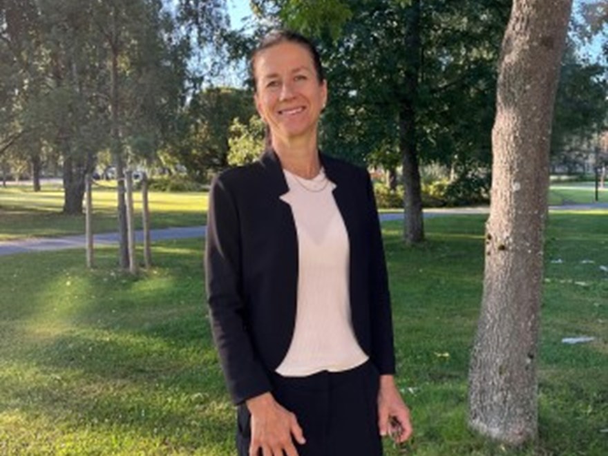 kvinna med mörkt hår i mörk jacka med rosa tröja står i träddunge