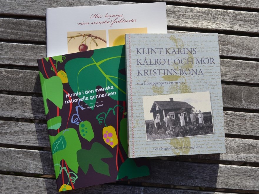 Färgfoto föreställande tre böcker som ligger på ett träbord. Böckerna är "Klint Karins kålrot och mor Kristins böna", "Humle i den svenska nationella genbanken" och "Här bevaras våra svenska fruktsorter".