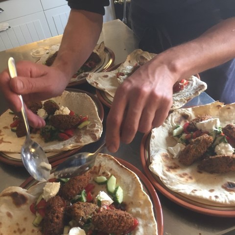 Färgfoto som föreställer tillagandet av falafel på gråärtor. På ett bord finns flera skålar med olika tillbehör och en person, som man bara ser händerna på, håller på att plocka ihop en falafelrulle.