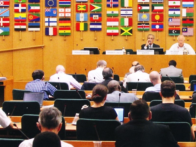 Färgfoto föreställande en grupp människor som sitter samlade i en stor möteslokal. Framför dem sitter flaggorna från alla FN:s medlemsländer uppsatta på väggen. 
