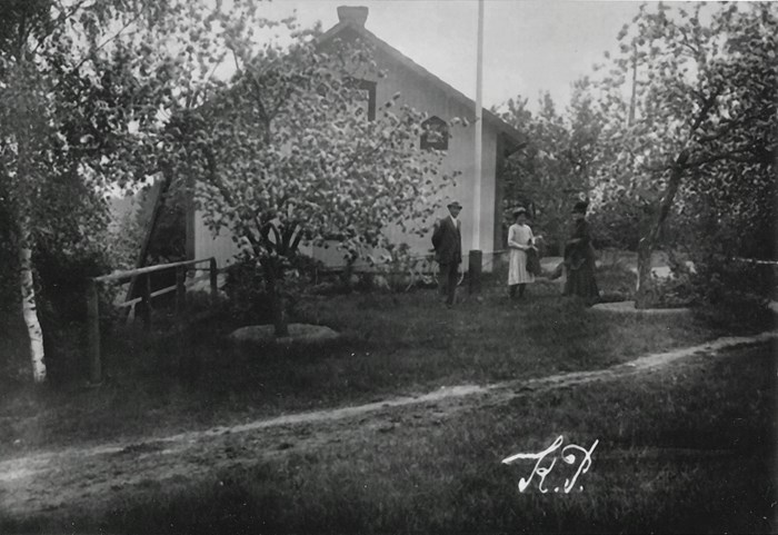 Ett svartvitt foto som föreställer tre personer som står i en trädgård framför gaveln på ett vitt trähus. 