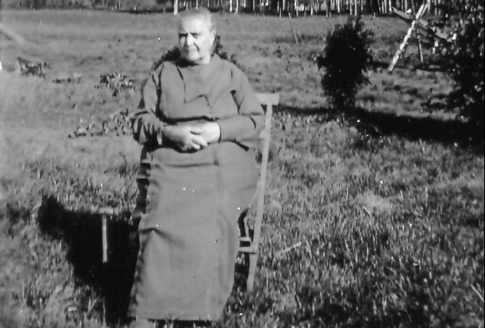 Svartvitt foto föreställande en äldre kvinna, Maria Eriksson, som sitter på en stol ute på en gräsmatta. I bakgrunden ses en åker och bakom den ett skogsparti.