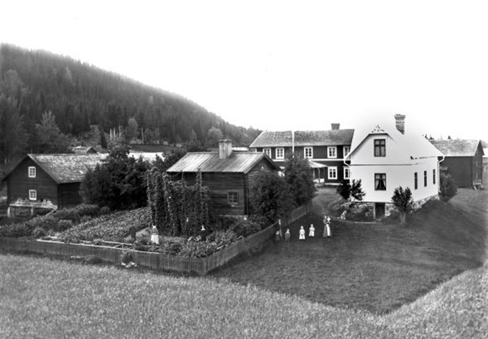 Svartvitt foto som visar en familj stående framför sin gård. Bostadshuset är ett  vitt trähus. Framför en av eknomibyggnaderna ligger en prydlig köksträdgård med flera humlestänger. 