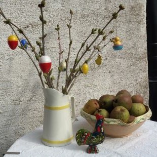 Färgfoto som visar ett påskris i en porslinskanna, ett keramikfat med päron och en svart tupp i trä. 