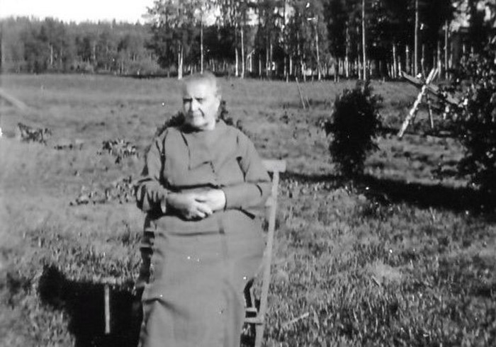 Ett svartvitt foto som visar en äldre kvinna som sitter på en stol utomhus. I bakgrunden ses ett öppet fält som ramas in av skog. 