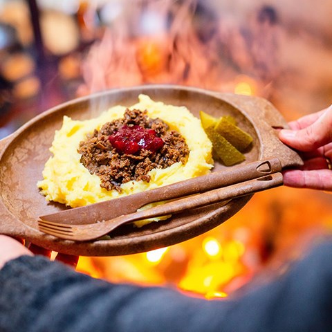Händer håller en tallrik med renskav och potatismos invid en öppen eld. Foto.