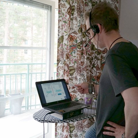 Hampus Holmström undervisar på distans. Ståendes framför ett fönster med datorn på en strykbräda och med ett headset.  