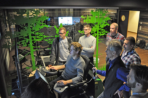 Sju personer står i ett rum och tittar på en demonstration av en skördarsimulator. Foto.