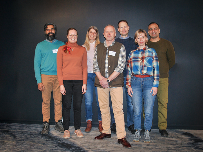 Seven people stands against a wall, looking happy. Photo.