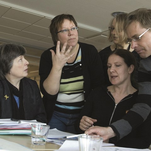 Fyra kvinnor och en man som sitter och en del står står kring ett bord och diskuterar något.