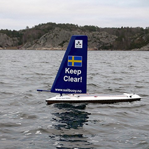 SLU Aqua Sailor in the water. It looks like a windsurfing board. On its sail is the SLU logo, a swedish flag and  the text Keep clear and www.sailbuoy.no.