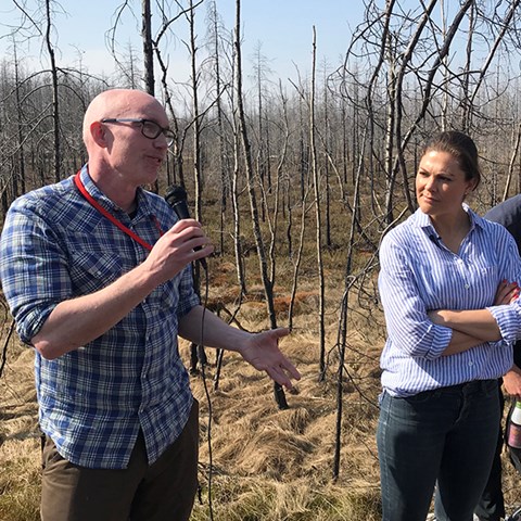 En forskare håller föredrag i bränd skog, i kanten syns en kvinna som lyssnar.