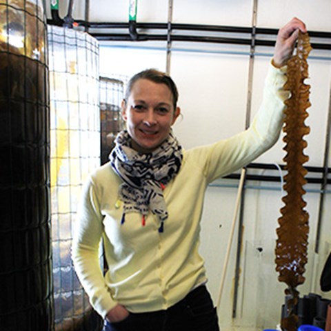 Närbild på Emma Ivarsson som håller upp en lång brunalg i vänster hand. Foto. 