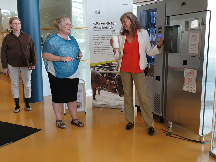Rektor Maria Knutson Wedel håller en mjölkflaska framför mjölkautomaten. Sigrid Agenäs och Rauni Niskanen står bredvid till vänster. Foto.