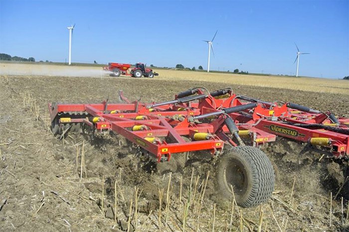 Ett jordbruksfält med ett rött jordbearbetningsredskap som kopplas till traktor. Foto.