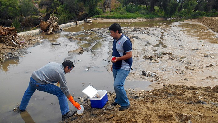 Två personer med handskar och vattenflaskor står och arbetar vid en flod. Foto.
