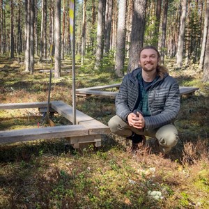 En man sitter hukad i skogen vid fyrkanter byggda av plankor. Foto.