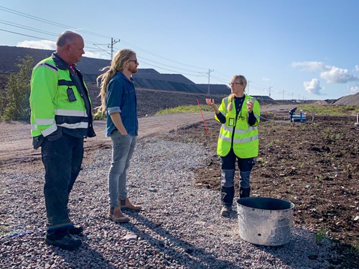 A woman talks to two men on gravelly ground in front of large piles of gravel. Photo.