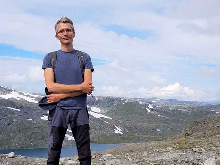 En man med korsade armar i friluftskläder. I bakgrunden syns fjäll. Foto.